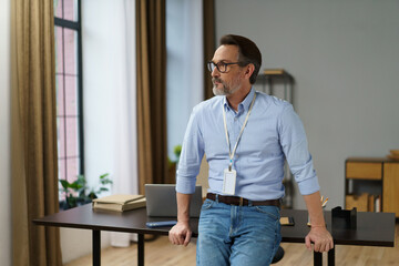 Fototapeta premium Man wearing a blue shirt and jeans is sitting at a desk. He has a name tag on and is looking at something
