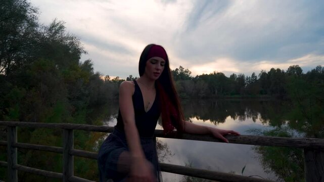 Chica joven realizando movimientos de danza contempor&aacute;nea con un paisaje natural de lago con bosque y el brillante y c&aacute;lido cielo del atardecer dibujando su silueta.