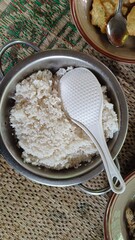 A bowl of freshly cooked white rice with a serving spoon on a table, with other plates and food in the background.