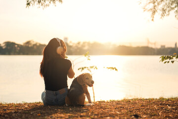 Happy Asian woman listen music in headphones relaxing with beagle dog pet and overlooks beautiful lake in outdoor sunset, Girls spend weekends in nature with a pet