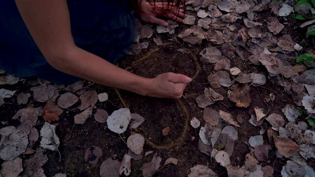 Chica joven sentada en un sendero en el bosque al atardecer, mira hacia abajo y mueve el brazo dibujando algo en el suelo. por mal de amores.