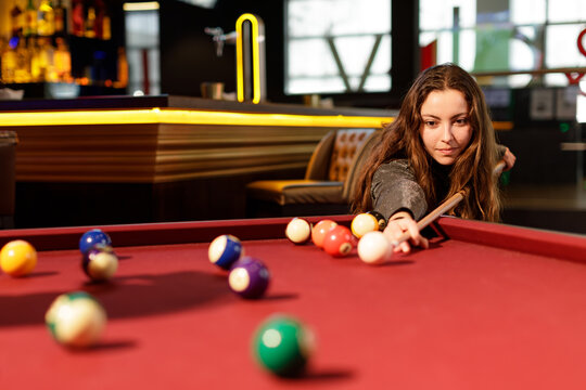 A concentrated young woman playing pool in a bar, prepares to accurately shoot the ball. Leisure and social environment concept.
