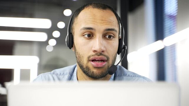 African American corporate operator who works in customer support speaks on a video call using a laptop. Call center agent in headset helping with complaints in office. Online consultation. Close up