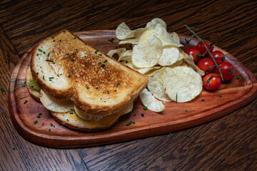 A sandwich with pickles potato chips on a plate with tomatoes on the side. The sandwich is served on a wooden plate.