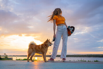 Young woman standing with her dog and skateboard by a river, soaking in the vibrant colors of a sunset, creating a tranquil and serene atmosphere filled with joy and freedom © wifesun