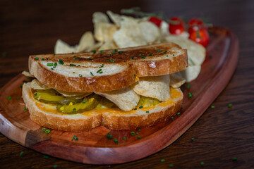 A sandwich with pickles potato chips on a plate with tomatoes on the side. The sandwich is served on a wooden plate.