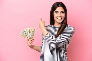 Young Brazilian woman taking a lot of money isolated on pink background pointing back