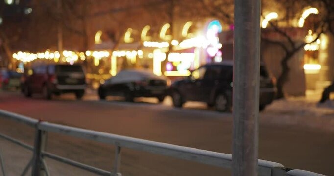 The seasons are winter. Cars with their headlights on drive along a snow-covered road past parked cars near a store decorated with glowing garlands.  Siberia.