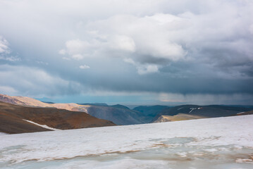 Dramatic alpine top view from flat sloping glacier on precipice edge to deep gorge between colorful sunlit cliffs and mountain range silhouette in rain under lead gray cloudy sky. Rainy grey clouds.