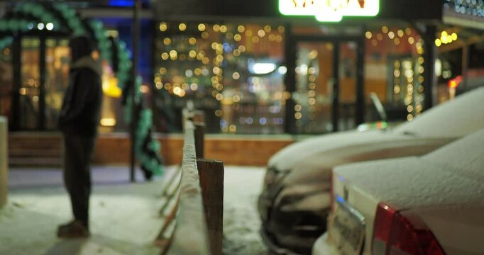 Night city. Novosibirsk.Out of focus. A man stands next to a parking lot of snow-covered cars against a background decorated with colorful garlands of storefronts. Hand held. Bokeh. Siberia.