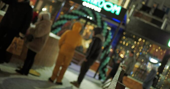 Holiday season. People are standing next to a parking lot of snow-covered cars against a backdrop of multi-colored garlands of storefronts decorated for Christmas. Hand held. Bokeh. Siberia.