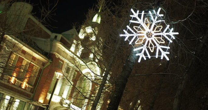 Holiday season, night city. A large snowflake made of lamps glows on a tree against the background of a beautifully illuminated building wall. The camera is rotating, hand held. Siberia.