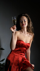 Happy beautiful young woman in festive red dress posing on camera, holding glass of champagne, getting ready for holiday. New Year concept. Merry Christmas