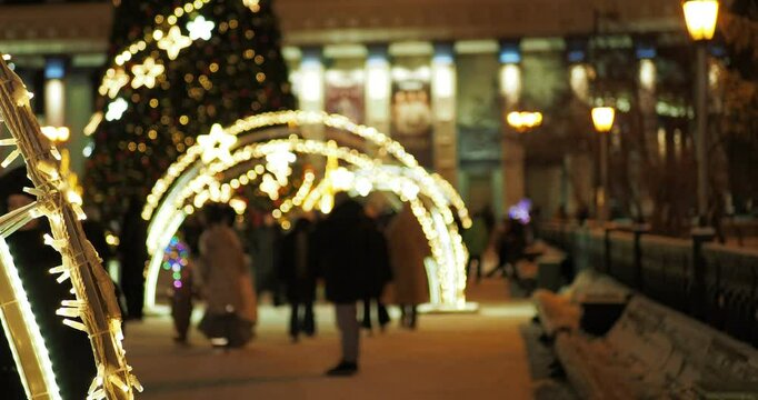 Holiday season. People are walking next to a Christmas tree and beautiful installations with iridescent multicolored lights against the background of a building with an illuminated facade. Hand held. 
