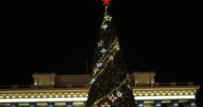 A Christmas tree with a red star on top is decorated with beautiful garlands of iridescent multicolored lights. The camera moves up. Hand held. Siberia.