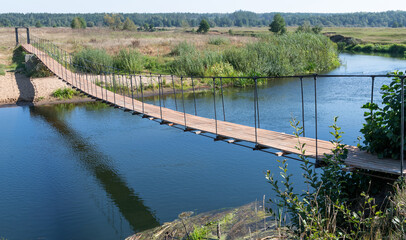 Unusual design. Suspended wooden bridge on steel cables or ropes. Not a wide river. Summer day. Forest, bushes, water, shadow and reflection of the bridge in the water.