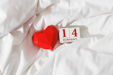 Valentine's Day concept, red paper valentine heart and 14 february holiday date on wooden calendar in bed on on  white crumpled sheets. Minimal style flat lay photo, top view, copy space