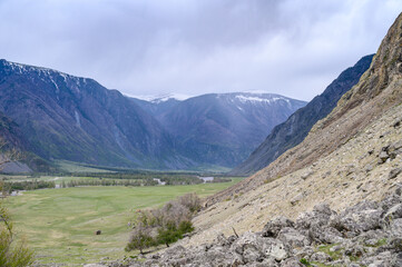 Fototapeta premium View from the top of a deep canyon surrounded by mountains, a river flowing below and a steep winding road descending down to the river on an overcast spring day. Chulyshman river valley, Altai,