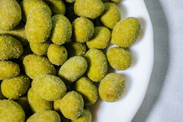 A close-up of a white plate filled with bright yellow-green wasabi-coated nuts. The nuts are coated in a fine powder, giving them a crunchy and spicy appeal.