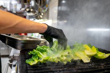 A chef's hand in black gloves carefully grills fresh romaine lettuce on a hot grill, surrounded by a steamy kitchen environment, showcasing vibrant green colors and culinary expertise.