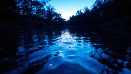 A calm river reflecting a deep blue sky at dusk , surrounded by silhouettes of trees on both sides. The water surface shows gentle ripples, creating a serene atmosphere .