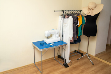 A sewing machine stands on a table with sentimental paper, chalk and pins, against the background of women's clothing and a black mannequin for cutting and sewing.