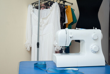 A sewing machine stands on a table with sentimental paper, chalk and pins, against the background of women's clothing and a black mannequin for cutting and sewing.