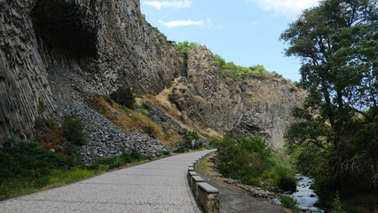 A winding pathway meanders along a rocky cliffside, surrounded by lush greenery and a gently flowing stream