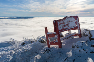 An old red bench on top of a mountain with a view of the mountain peaks above the clouds.