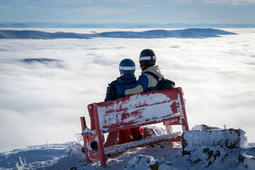 Couple of skiers on a bench on top of a mountain above the clouds. Man and woman with ski helmets admiring beautiful winter landscape in the mountains sitting on the peak of the mountain.