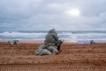 A tired man is sitting on a sack by the sea, dressed in a protective suit. Puts fuel oil in a bag....