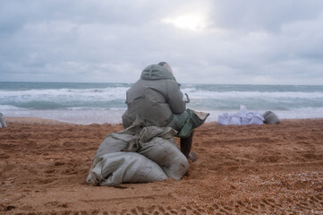 A tired man is sitting on a sack by the sea, dressed in a protective suit. Puts fuel oil in a bag....