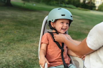 A parent fastens their child's helmet strap while the child laughs in a rear bike seat, ensuring...