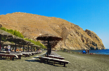 Popular Perissa beach with sun lounger and straw umbrellas on black volcanic sand near rock Mesa Vouno.