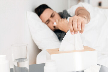Fototapeta premium Sick young man lying in bed and sneezing runny nose, taking napkins, selective focus on tissue box on bedside table, bedroom interior, copy space. Cold, flue, coronavirus, COVID-19 concept