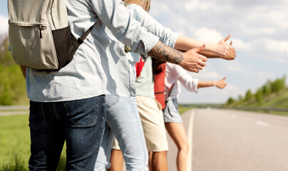 Obraz premium Group of unrecognizable young friends standing along road, making thumb up gesture, hitchhiking for car during road trip, traveling together in summer, closeup. Outdoor vacation, tourism concept