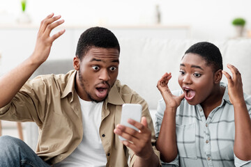Shocked African American Tourists Couple With Smartphone Reading Shocking Travel News About Tourism Sitting Near Unpacked Suitcase At Home. Traveling App Advertisement Concept