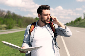 Handsome young man standing on road with map, feeling lost, calling on smartphone, checking travel options, empty space. Stressed millennial guy hitchhiking on highway, cannot catch ride