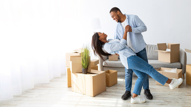 Happy loving african american couple dancing in their new house among carton boxes on moving day, panorama. Excited young family having fun while relocating to new property - Powered by Adobe