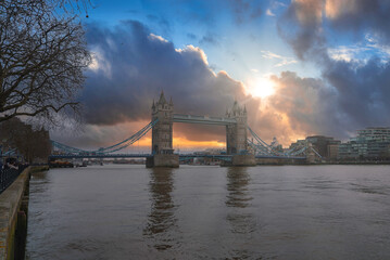 Tower Bridge stands over the River Thames in London, with the sun breaking through clouds. Bare...