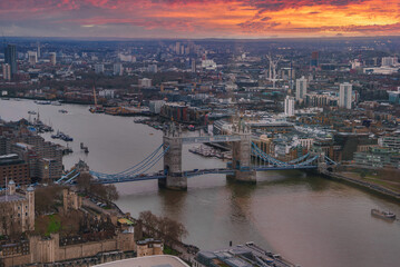 View of Tower Bridge spanning the River Thames in London at sunset. The sky is painted in orange and red hues, reflecting on the river below.