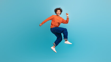 A young African American man with curly hair is jumping joyfully against a vibrant blue background. He is wearing a casual orange sweater and blue jeans, exuding youthful energy and enthusiasm.
