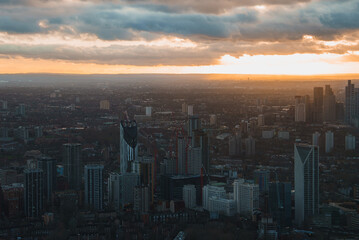 Fototapeta premium Aerial view of London during sunset, showcasing modern skyscrapers like Strata SE1. The cityscape is illuminated by a warm glow against dark clouds.