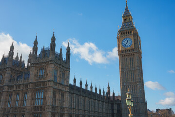 Obraz premium The iconic Big Ben and Gothic architecture of the Houses of Parliament stand prominently under a clear blue sky with scattered clouds in London, England.