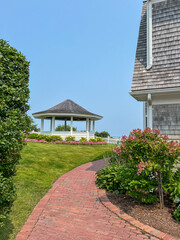 A brick path leads to a gazebo surrounded by lush green grass and pink blooming flowers, with a clear blue sky in the background and a coastal view peeking out in the distance.