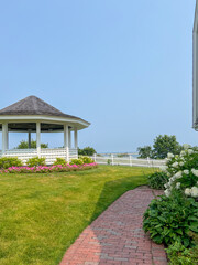 A charming brick path leads toward a gazebo surrounded by colorful flowers and lush greenery, with a white picket fence and serene ocean views in the distance under a clear blue sky.