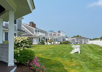 A sunny lawn with white Adirondack chairs and blooming hydrangeas leading to a beach house with a fence in the background, perfect for relaxing and enjoying a coastal view.