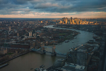 Aerial view of London featuring Tower Bridge over the River Thames, Canary Wharf skyscrapers, and City Hall, all illuminated by warm, golden light.