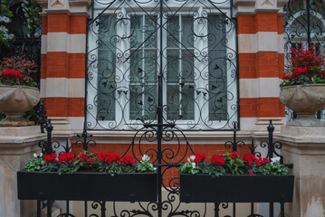 An intricately designed wrought iron gate stands before a red and cream brick building. White framed windows and vibrant flowers suggest a Christmas theme.