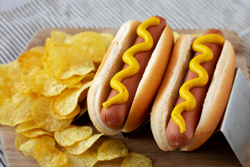 Homemade Boiled Hot Dogs with Mustard and Potato Chips on a Wooden Board, side view.